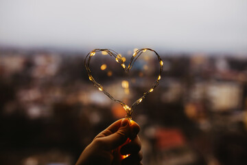 Hand holding heart made of fairy lights on window background.
