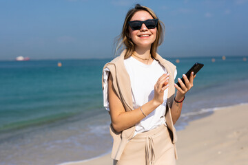 Young stylish woman in a summer outfit, white shirt use mobile phone on the beach. Happy attractive traveler woman looks at the screen of gadget at vacations.