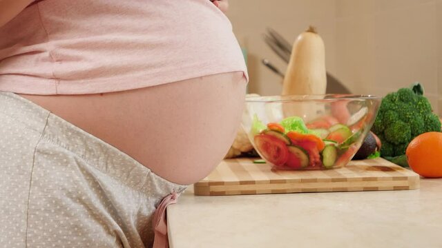 Closeup of pregnant woman with big belly standing on kitchen and drinking water from glass. Concept of healthy lifestyle, nutrition and hydration during pregnancy