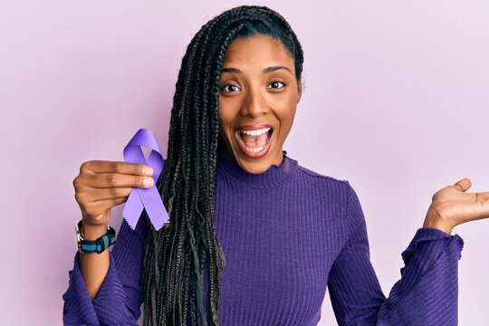 African american woman holding purple ribbon awareness celebrating achievement with happy smile and winner expression with raised hand
