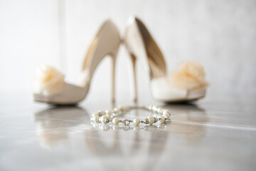 Pearl bracet in front of High heel bridal shoes with silk flowers on reflective surface