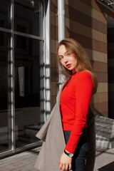 Portrait of a young woman taking off coat, looking at camera and standing on street in front of window and brown wall. Caucasian girl having fun outside. Urban style and fashion. Casual outfit