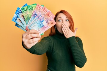 Beautiful redhead woman holding swiss franc banknotes covering mouth with hand, shocked and afraid for mistake. surprised expression