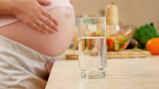 Closeup of full glass of water against pregnant woman touching and caressing big belly on kitchen. Concept of healthy lifestyle, nutrition and hydration during pregnancy
