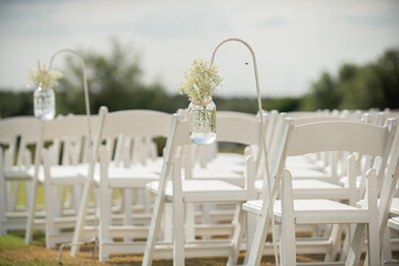 Fancy white folding chairs lined  up at outdoor wedding ceremony with babyie's breathe flowers in mason jars hanging