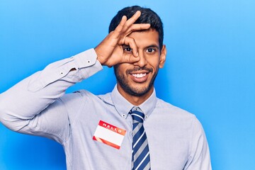 Young latin man wearing sticker with hello my name is message smiling happy doing ok sign with hand on eye looking through fingers