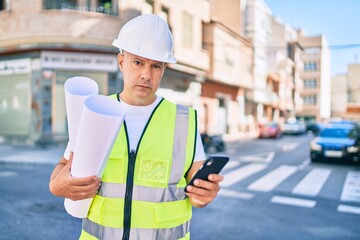 Middle age architect man with serious expression using smartphone and holding blueprints at the city.