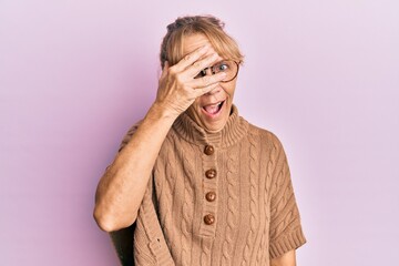Middle age blonde woman wearing glasses over pink background peeking in shock covering face and eyes with hand, looking through fingers afraid