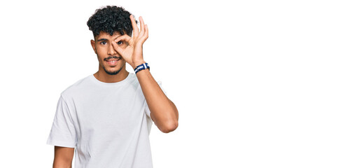 Young arab man wearing casual white t shirt doing ok gesture with hand smiling, eye looking through fingers with happy face.