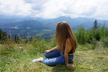 A girl with dark hair, in jeans and a black T-shirt, sits on a lawn and looks at the mountains. view from the back. without a face.