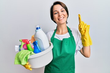 Young brunette woman with short hair wearing apron holding cleaning products smiling amazed and surprised and pointing up with fingers and raised arms.