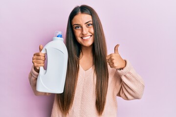 Beautiful hispanic woman holding laundry detergent bottle smiling happy and positive, thumb up...