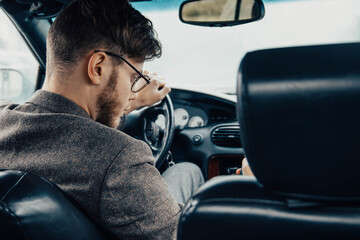 male car driver in glasses and a jacket drives a car during the day.