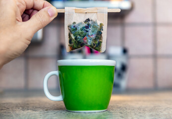 Hand holding a filter bag with wooden stick filled with herbal tea above a cup, kitchen background.