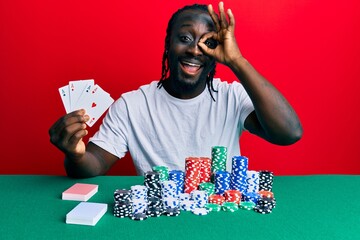 Handsome young black man playing poker holding cards smiling happy doing ok sign with hand on eye looking through fingers