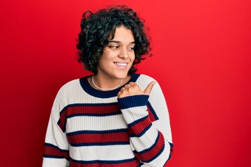 Fototapeta na wymiar Young hispanic woman with curly hair wearing casual winter sweater pointing thumb up to the side smiling happy with open mouth
