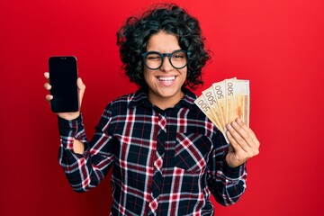 Young hispanic woman with curly hair showing smartphone screen holding 500 norwegian banknotes...
