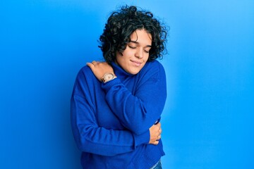 Fototapeta na wymiar Young hispanic woman with curly hair wearing casual winter sweater hugging oneself happy and positive, smiling confident. self love and self care