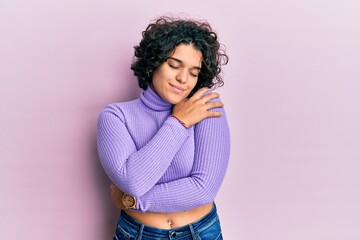 Young hispanic woman with curly hair wearing turtleneck sweater hugging oneself happy and positive, smiling confident. self love and self care