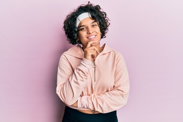 Fototapeta na wymiar Young hispanic woman with curly hair wearing sportswear looking confident at the camera with smile with crossed arms and hand raised on chin. thinking positive.