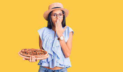 Young beautiful latin girl wearing summer hat and glasses holding pizza covering mouth with hand,...