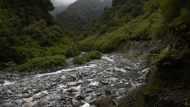 Elavated view of a fast-flowing, mountain stream flowing along a boulder-strewn valley floor and between forested walls. Tatare Stream, Westland Tai Poutini National Park, New Zealand.