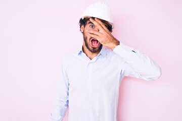 Handsome young man with curly hair and bear wearing architect hardhat peeking in shock covering face and eyes with hand, looking through fingers afraid