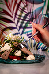 A feminine hand tasting an Aesthetic Tomato Salad with tuna, Rocket and Mozzarella. Modern Spanish cuisine salad.The background is full of colored plants.