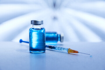 A vial of blue liquid vaccine lays on a metal medical surface next to a syringe ready for treatment