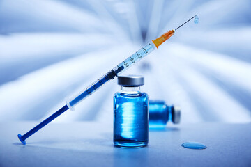 A syringe with blue liquid vaccine leans against a vial holding treatment on a metal medical surface