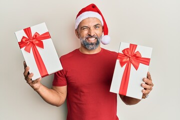 Middle age handsome man wearing christmas hat and holding a gifts winking looking at the camera...