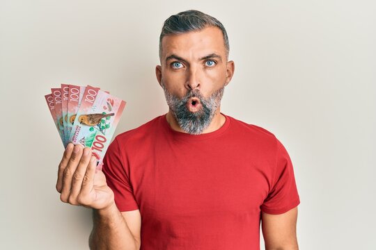 Middle age handsome man holding 100 new zealand dollars banknote scared and amazed with open mouth for surprise, disbelief face