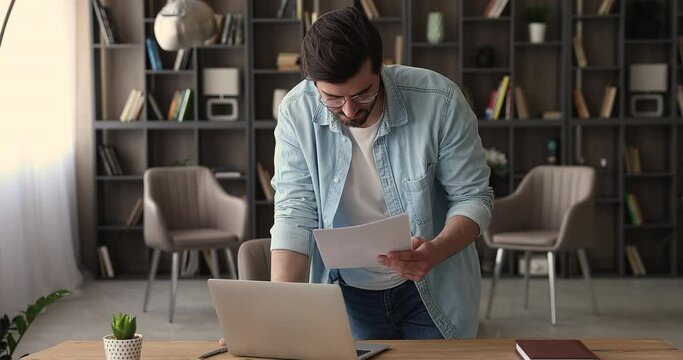 Man standing in office room lean over workplace desk hold documents work on laptop, analyzing company financial paperwork, thinking, consider statistics papers, preparing report, make research concept