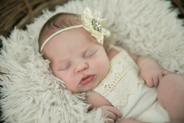 Close up of a sleeping sweet newborn infant baby girl with a cream-colored bonnet hat on laying on a pale neutral background laying in a basket wreath