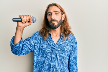 Attractive man with long hair and beard singing song using microphone thinking attitude and sober expression looking self confident