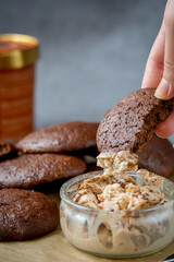 Woman's hand picking up a fluffy chocolate brownie cookie dipped in a glass bowl of peanut butter ice cream. Healthy homemade gluten free chocolate chip cookies in the background with ice cream bucket