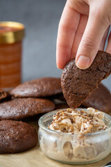 Woman's hand wanting to dip a fluffy chocolate brownie cookie in a glass bowl of peanut butter ice cream. Healthy homemade gluten-free chocolate chip cookies in the background with ice cream bucket