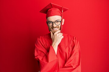 Young redhead man wearing red graduation cap and ceremony robe looking confident at the camera smiling with crossed arms and hand raised on chin. thinking positive.