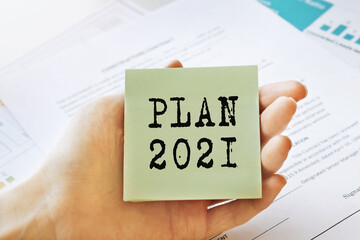 Close-up female hands hold a sign with the inscription PLAN 2021 on the background of business charts and business documents. Close-up, soft light. Marketing concept.