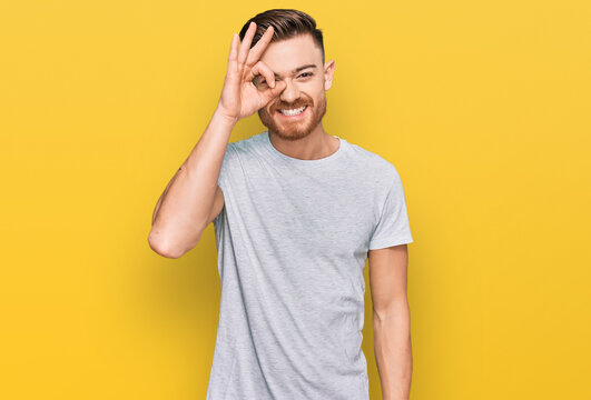 Young redhead man wearing casual grey t shirt doing ok gesture with hand smiling, eye looking through fingers with happy face.