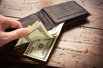 Hand taking out money from one hundred US banknotes in leather wallet on wooden table background. Cash of hundred dollar bills, paper money currency.