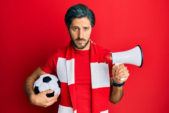 Young hispanic man football hooligan holding ball using megaphone depressed and worry for distress, crying angry and afraid. sad expression.