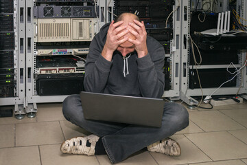 A sad technician put his hands on his head in the server room. Man with a laptop sits on the floor...