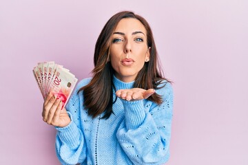 Young brunette woman holding 20 israel shekels banknotes looking at the camera blowing a kiss with hand on air being lovely and sexy. love expression.