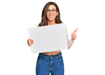 Fototapeta premium Young brunette woman holding blank empty banner smiling happy and positive, thumb up doing excellent and approval sign