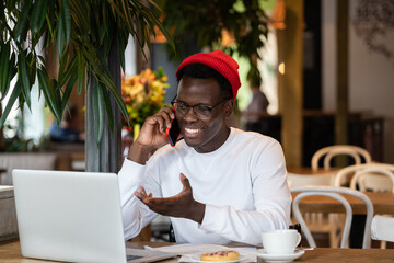 Happy millennial Afro-American hipster man wear red hat laughing, speaking with friend on mobile phone, enjoying watching educational webinar on laptop, remotely online work in cafe during lunchtime