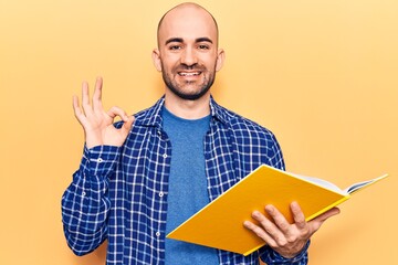 Young handsome bald man reading book doing ok sign with fingers, smiling friendly gesturing excellent symbol