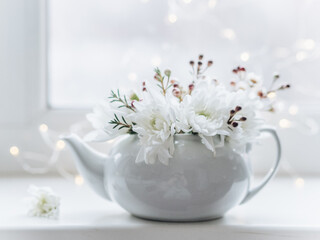 A white ceramic teapot for making tea stands on the windowsill. There are white chrysanthemums in the teapot. Spring drink. Spring came. Flower tea. Romantic mood. Unusual flower vase. Spring flowers.