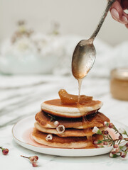 American pancakes are decorated with white flowers. On top of the pancakes, pour berry jam from a spoon. Breakfast for Valentine's Day. Pancakes with jam. Decorate food with flowers.