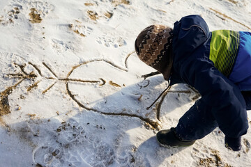 Little boy draws with enthusiasm a big heart with an arrow on the snowy sand.